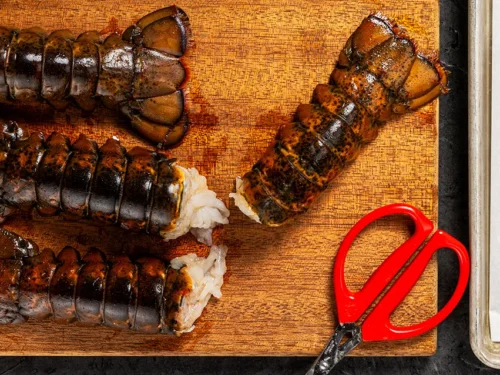 Frozen lobster tails on a wooden cutting board with kitchen shears, prepared for butterflying, next to a baking sheet with split lobster tails ready for cooking.